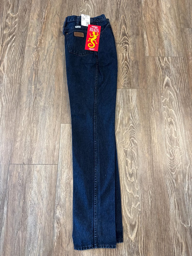 May include: Dark blue denim jeans with a brown leather patch and a red tag with the word "RIATA" and a yellow lizard graphic. The jeans are folded and lying on a wooden surface.