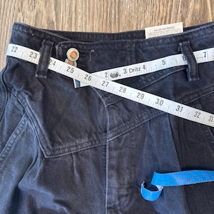May include: Close-up of black denim trousers with a distinctive design. A white measuring tape is wrapped around the waist, showing measurements. The trousers have a button closure and a small tag. A blue measuring tape is also visible.