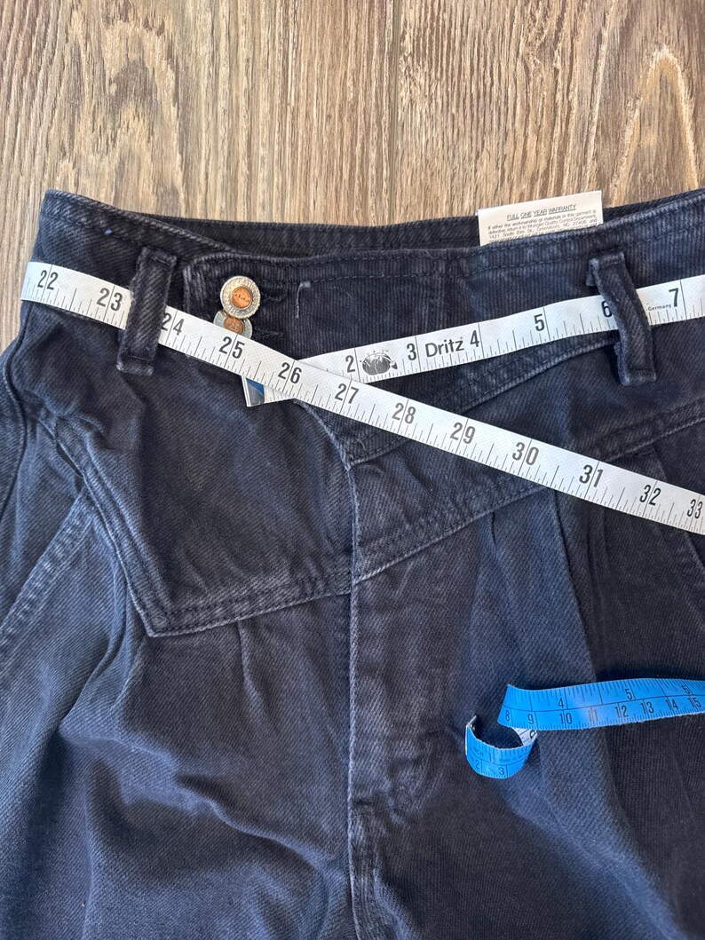 May include: Close-up of black denim trousers with a distinctive design. A white measuring tape is wrapped around the waist, showing measurements. The trousers have a button closure and a small tag. A blue measuring tape is also visible.