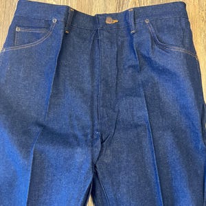 May include: Close-up of dark blue denim jeans. The jeans have a button closure, front pockets, and a classic design. The stitching is a contrasting tan color.