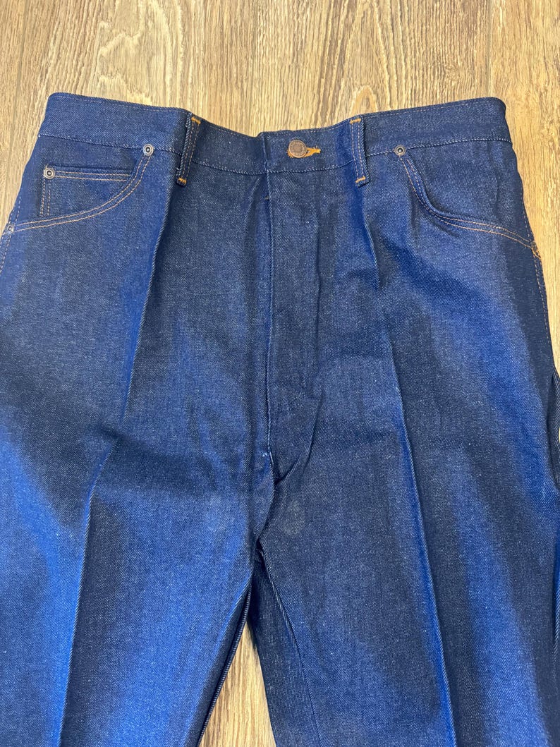 May include: Close-up of dark blue denim jeans. The jeans have a button closure, front pockets, and a classic design. The stitching is a contrasting tan color.