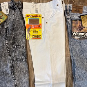 K&ouml;nnte beinhalten: Drei Wrangler-Jeans in verschiedenen Waschungen. Eine ist s&auml;uregewaschen, eine ist wei&szlig; und eine ist mittelblau. Jede Hose hat Etiketten. Die wei&szlig;e Hose hat ein "ProRodeo Cowboy Cut"-Etikett.