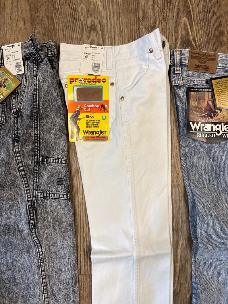 K&ouml;nnte beinhalten: Drei Wrangler-Jeans in verschiedenen Waschungen. Eine ist s&auml;uregewaschen, eine ist wei&szlig; und eine ist mittelblau. Jede Hose hat Etiketten. Die wei&szlig;e Hose hat ein "ProRodeo Cowboy Cut"-Etikett.