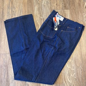 May include: Dark blue denim jeans with a straight leg design. The jeans have a classic five-pocket style and a Wrangler tag attached. The stitching is a contrasting tan color.