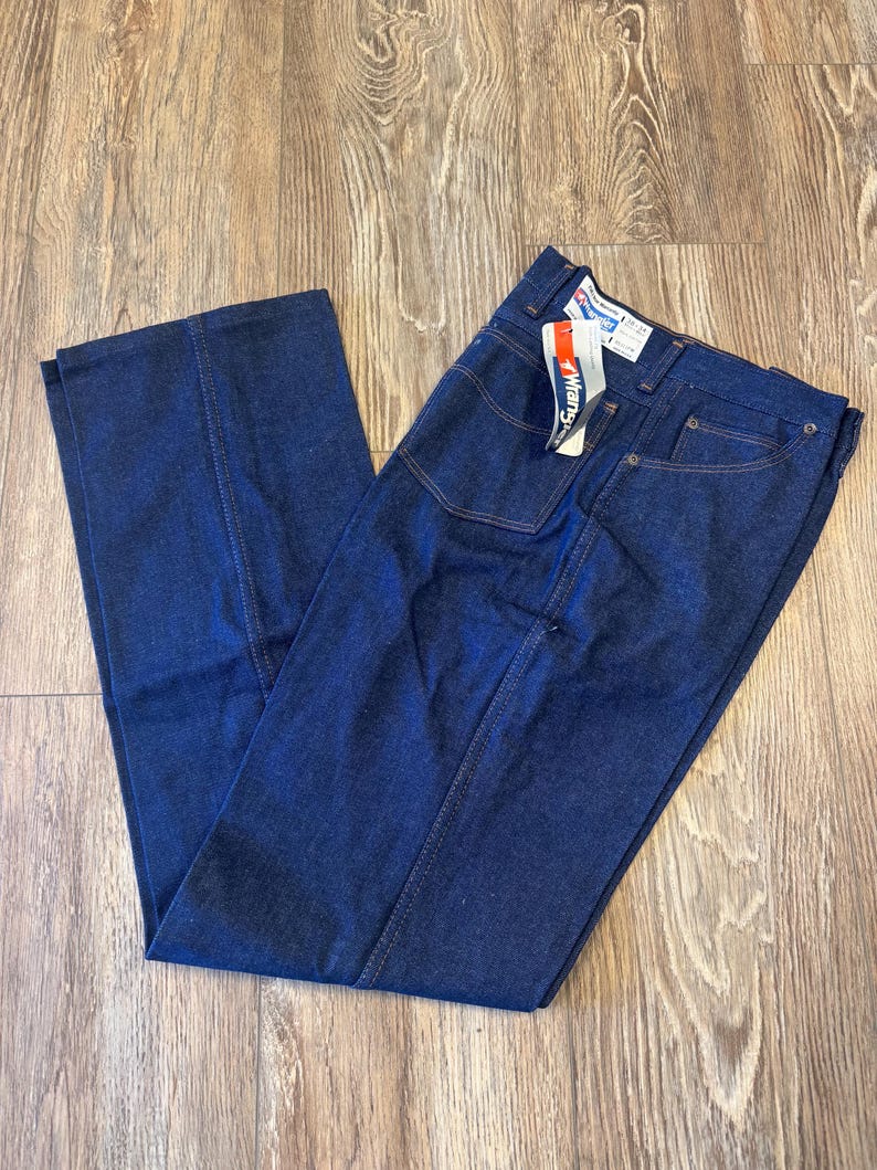 May include: Dark blue denim jeans with a straight leg design. The jeans have a classic five-pocket style and a Wrangler tag attached. The stitching is a contrasting tan color.