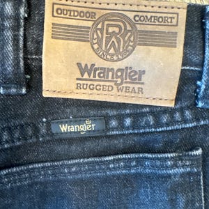 Puede incluir: Primer plano de un par de pantalones vaqueros Wrangler negros con un parche de cuero marr&oacute;n que dice "Outdoor Comfort" y "Wrangler Rugged Wear". El parche tambi&eacute;n presenta un logotipo con las letras "R" y "W" dentro de un c&iacute;rculo con el texto "Since 1904".