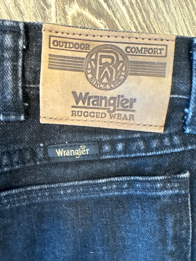Puede incluir: Primer plano de un par de pantalones vaqueros Wrangler negros con un parche de cuero marr&oacute;n que dice "Outdoor Comfort" y "Wrangler Rugged Wear". El parche tambi&eacute;n presenta un logotipo con las letras "R" y "W" dentro de un c&iacute;rculo con el texto "Since 1904".