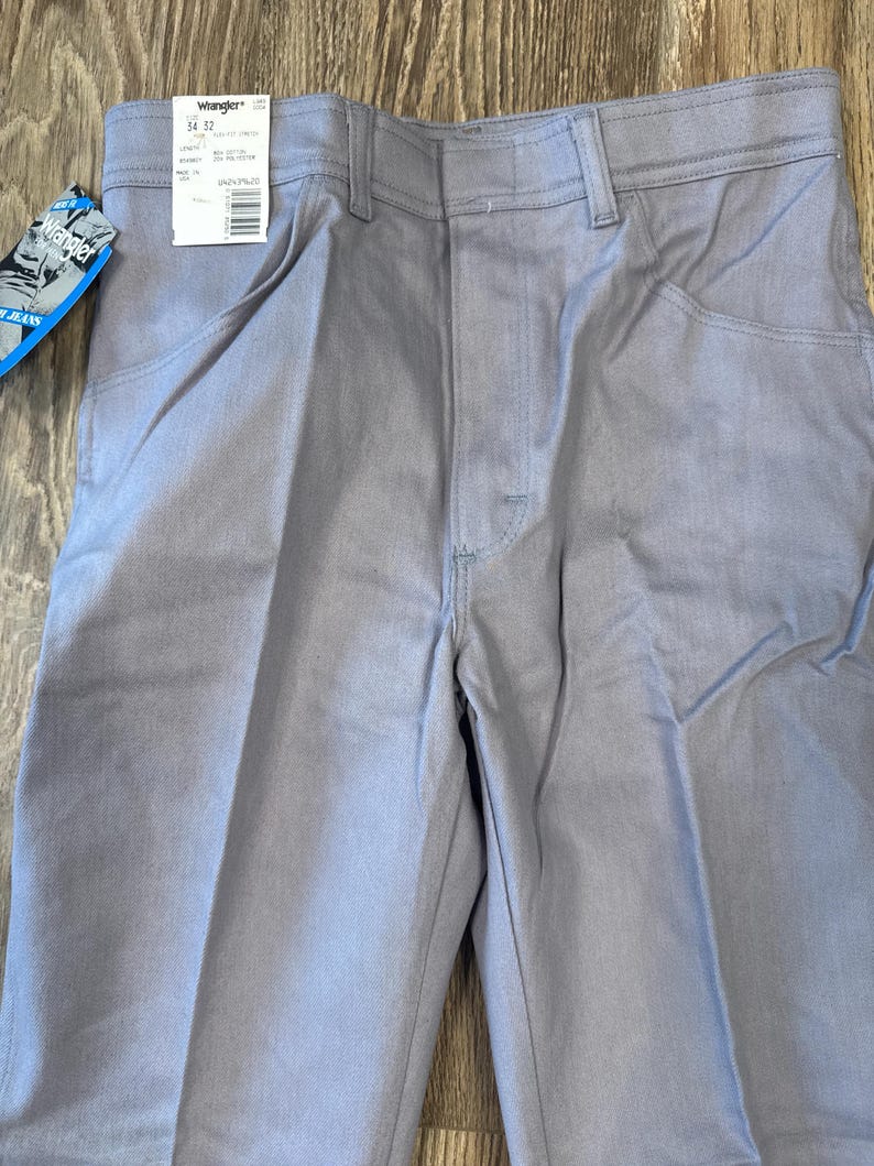 May include: Light gray Wrangler jeans with a tag attached. The tag displays the brand name and size information. The jeans have a classic five-pocket design and a straight leg cut. The fabric appears to be a durable denim material.
