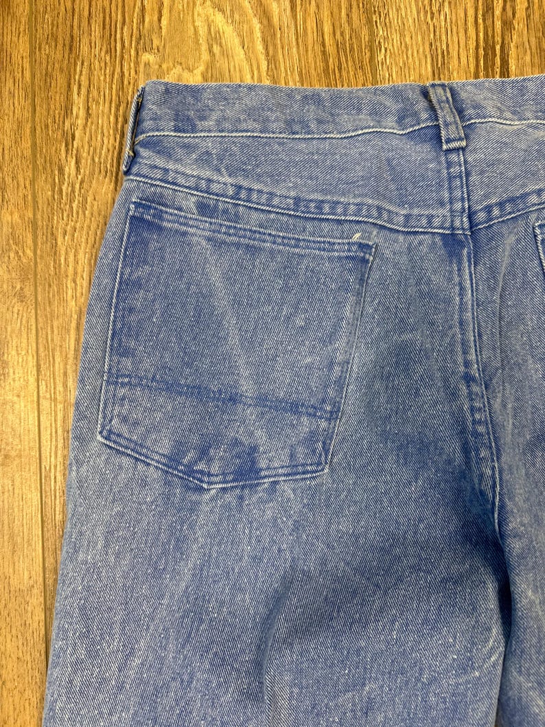 May include: Close-up of the back of light blue denim jeans. The jeans have a single back pocket and a waistband with belt loops. The denim has a faded appearance, with lighter areas creating a worn look. The stitching is visible along the seams and pocket edges.