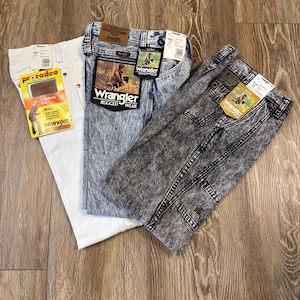 K&ouml;nnte beinhalten: Drei Wrangler-Jeans in verschiedenen Waschungen: Wei&szlig;, Acid-Wash-Blau und Grau. Jede Hose hat Etiketten, mit dem Wrangler-Logo und dem Text "Rugged Wear" auf der blauen Hose. Die wei&szlig;e Hose hat ein "prorodeo"-Etikett.