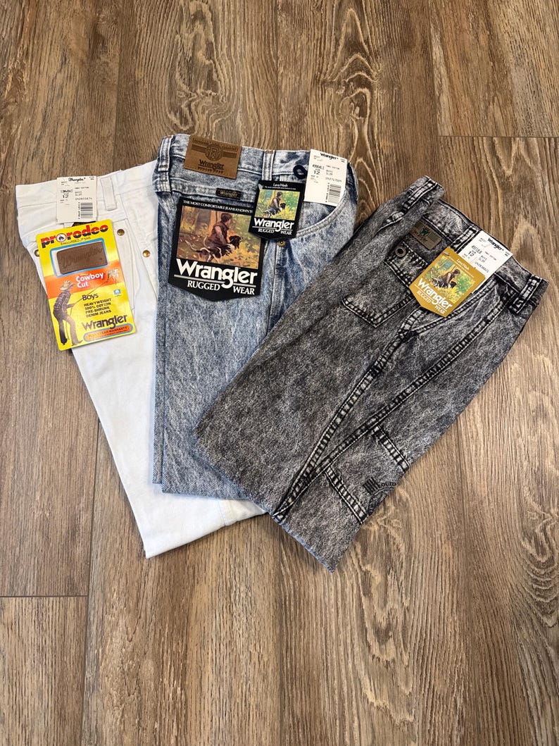 K&ouml;nnte beinhalten: Drei Wrangler-Jeans in verschiedenen Waschungen: Wei&szlig;, Acid-Wash-Blau und Grau. Jede Hose hat Etiketten, mit dem Wrangler-Logo und dem Text "Rugged Wear" auf der blauen Hose. Die wei&szlig;e Hose hat ein "prorodeo"-Etikett.