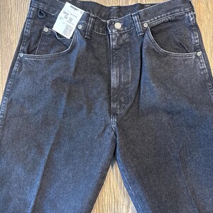 Puede incluir: Un par de pantalones vaqueros negros de mezclilla con una etiqueta Wrangler. La etiqueta dice "Talla 34" y "Cintura 32".