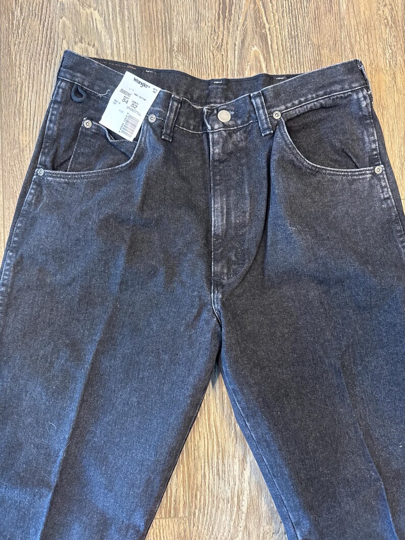 Puede incluir: Un par de pantalones vaqueros negros de mezclilla con una etiqueta Wrangler. La etiqueta dice "Talla 34" y "Cintura 32".