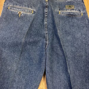 May include: Close-up of the back of blue denim jeans. The jeans have two back pockets with yellow stitching and a small green and grey Wrangler label. The jeans are made of a medium-wash denim material.
