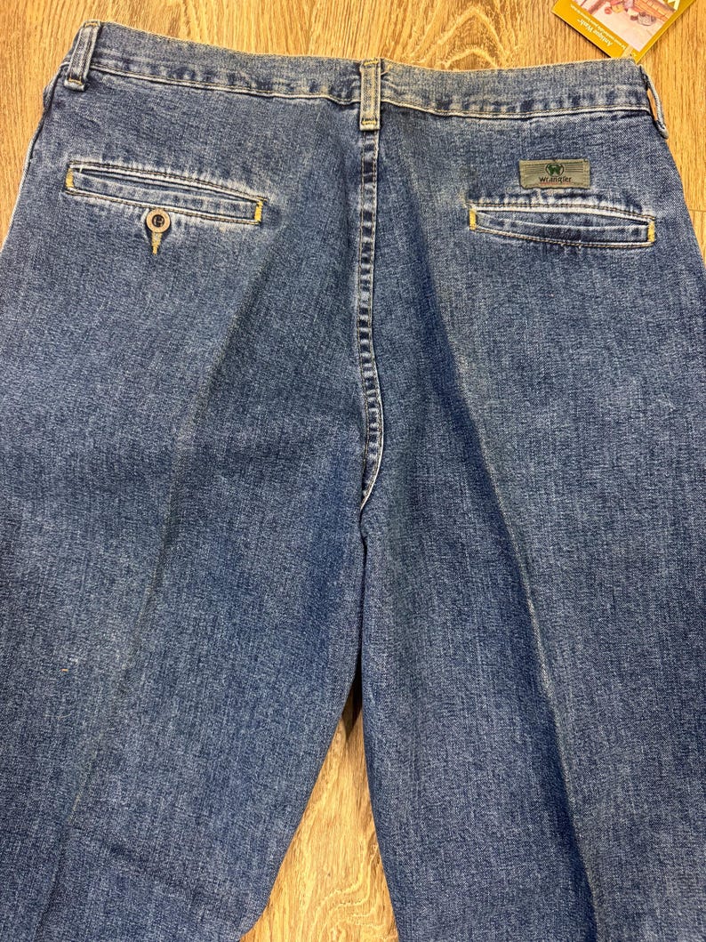 May include: Close-up of the back of blue denim jeans. The jeans have two back pockets with yellow stitching and a small green and grey Wrangler label. The jeans are made of a medium-wash denim material.