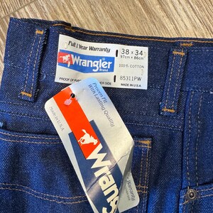 May include: Close-up of dark blue Wrangler jeans with a tag. The jeans have a 38x34 size label, 100% cotton, and a "Full 1 Year Warranty" label. The tag reads "Proven Fit With Lasting Quality".
