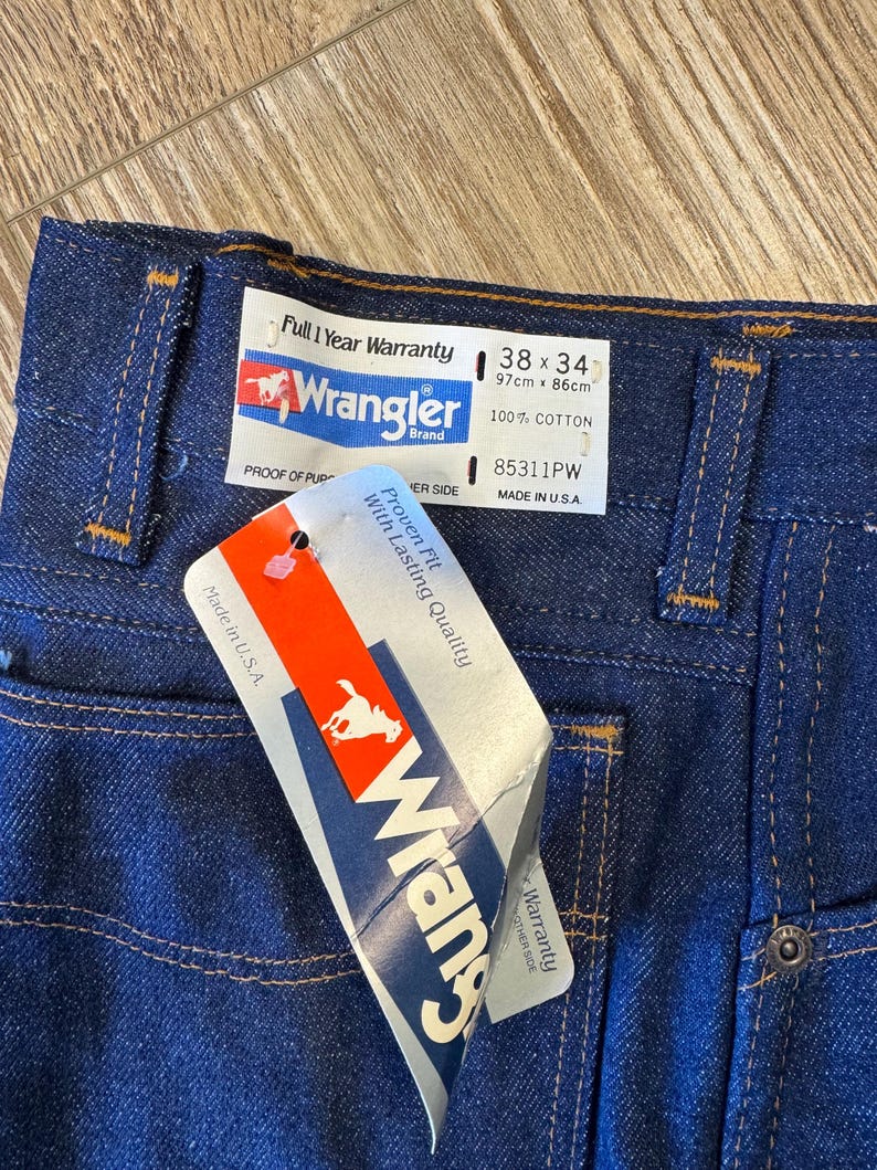 May include: Close-up of dark blue Wrangler jeans with a tag. The jeans have a 38x34 size label, 100% cotton, and a "Full 1 Year Warranty" label. The tag reads "Proven Fit With Lasting Quality".