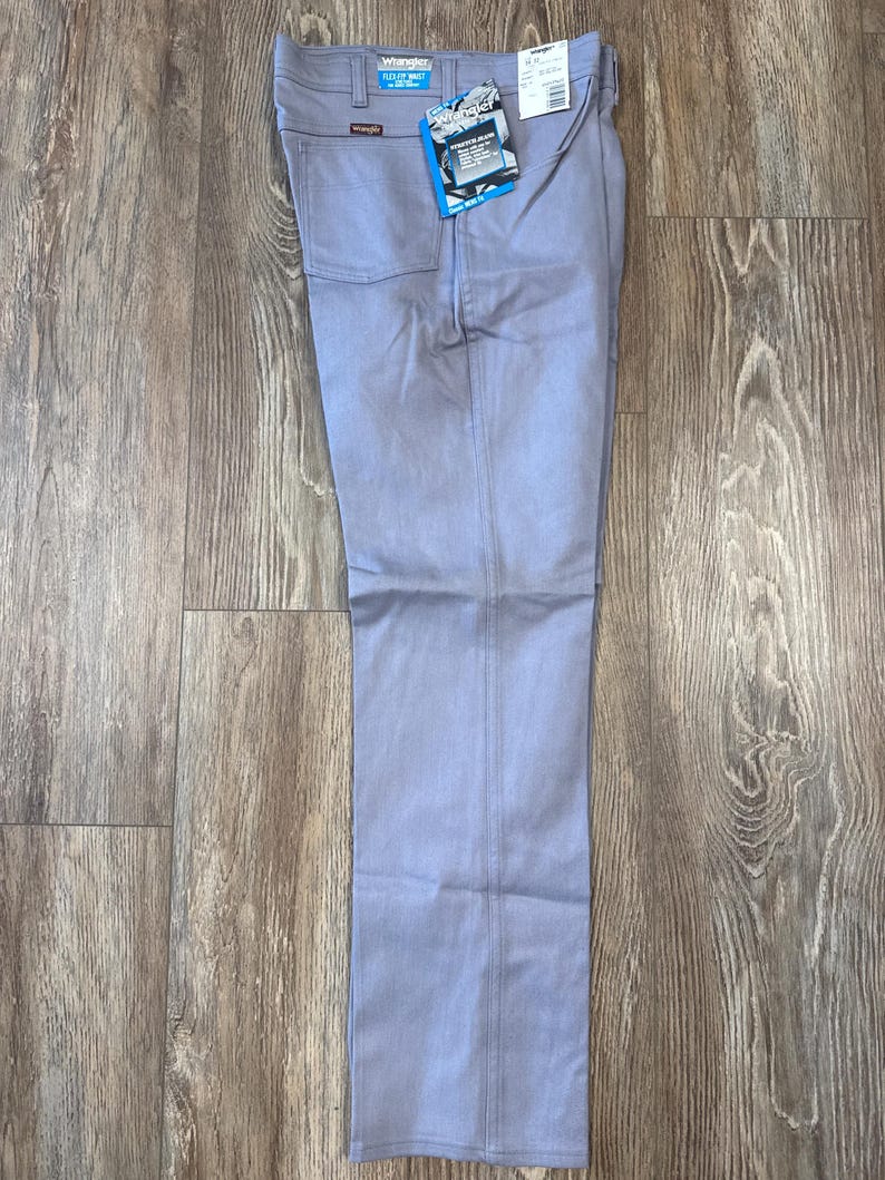 May include: Light blue Wrangler Flex Fit jeans with a straight leg. The jeans have a classic five-pocket design and a tag attached to the front. The jeans are laid flat on a wooden surface.