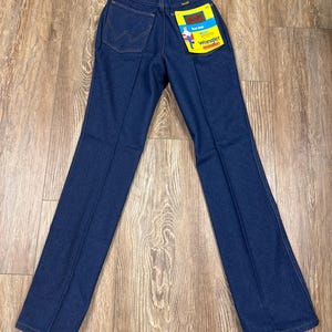 K&ouml;nnte beinhalten: Dunkelblaue Wrangler-Jeans mit klassischer Passform. Die Jeans hat ein gerades Bein und eine hohe Taille. Die Ges&auml;&szlig;tasche zeigt das Wrangler-Logo. Ein gelbes Etikett mit dem Wrangler-Logo und Text ist an der Ges&auml;&szlig;tasche befestigt.