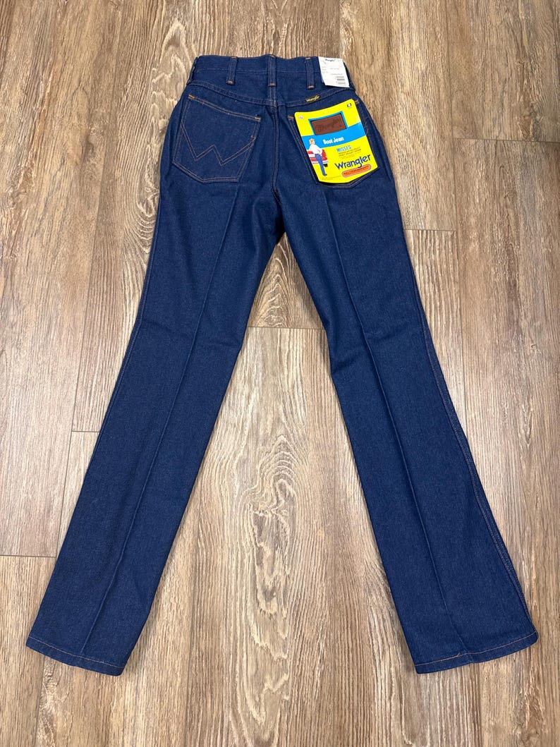 K&ouml;nnte beinhalten: Dunkelblaue Wrangler-Jeans mit klassischer Passform. Die Jeans hat ein gerades Bein und eine hohe Taille. Die Ges&auml;&szlig;tasche zeigt das Wrangler-Logo. Ein gelbes Etikett mit dem Wrangler-Logo und Text ist an der Ges&auml;&szlig;tasche befestigt.