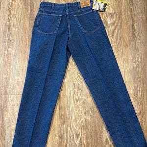 May include: Dark blue denim jeans with a classic five-pocket design. The jeans have a straight leg cut and a Wrangler brand tag. The back pockets are square. The jeans are laid flat on a wooden surface.