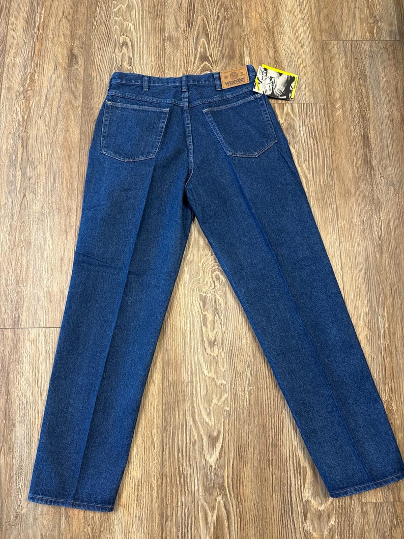 May include: Dark blue denim jeans with a classic five-pocket design. The jeans have a straight leg cut and a Wrangler brand tag. The back pockets are square. The jeans are laid flat on a wooden surface.