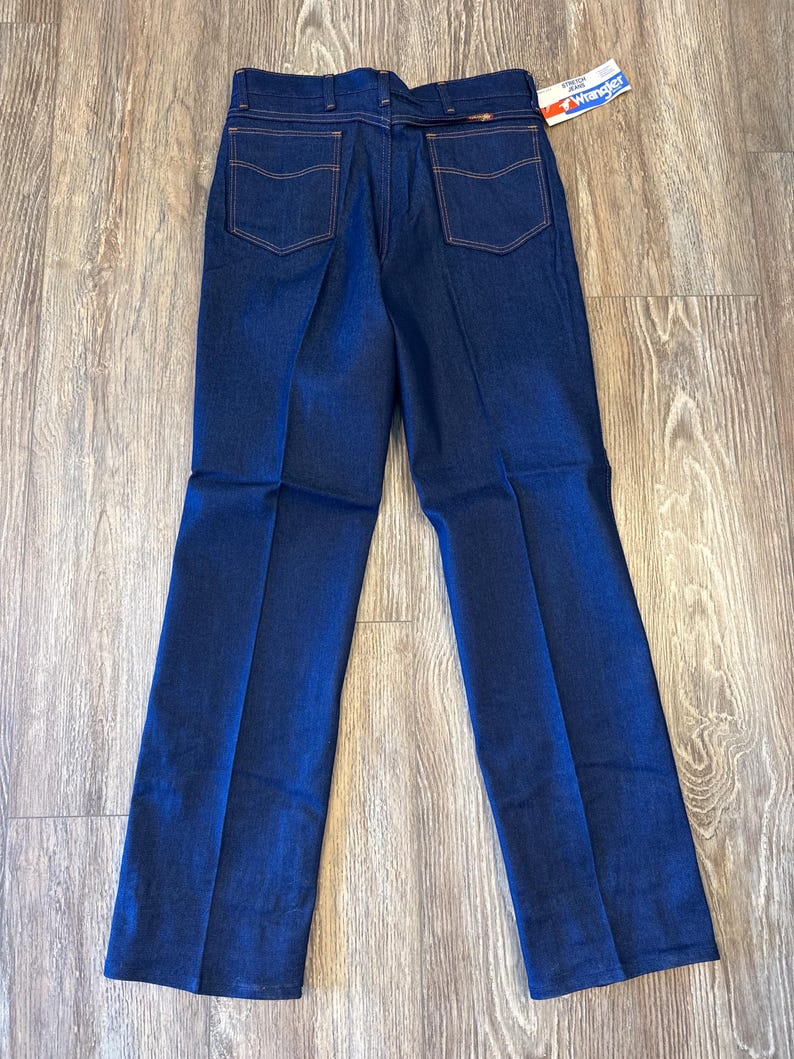 Peut inclure: Un jean en denim bleu fonc&eacute; &agrave; jambe droite. Le jean a un &eacute;cusson en cuir marron sur la ceinture arri&egrave;re avec le mot "Wrangler" imprim&eacute; dessus. Le jean a une &eacute;tiquette attach&eacute;e &agrave; la ceinture arri&egrave;re qui dit "Genuine Western Jeans" et "Wrangler".
