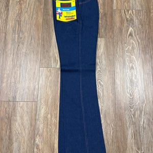 K&ouml;nnte beinhalten: Dunkelblaue Wrangler Bootcut-Jeans mit gelb-blauem Etikett. Die Jeans haben ein klassisches F&uuml;nf-Taschen-Design und sind aus strapazierf&auml;higem Denim-Stoff gefertigt. Das Etikett zeigt das Wrangler-Logo und eine Grafik eines Cowboys.