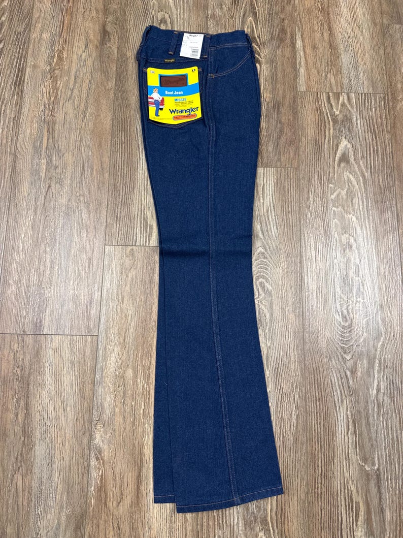 K&ouml;nnte beinhalten: Dunkelblaue Wrangler Bootcut-Jeans mit gelb-blauem Etikett. Die Jeans haben ein klassisches F&uuml;nf-Taschen-Design und sind aus strapazierf&auml;higem Denim-Stoff gefertigt. Das Etikett zeigt das Wrangler-Logo und eine Grafik eines Cowboys.
