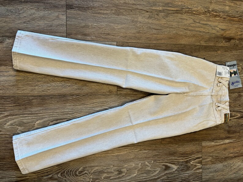 May include: A pair of white striped Wrangler jeans with a size tag attached. The jeans are laid flat on a wooden surface.