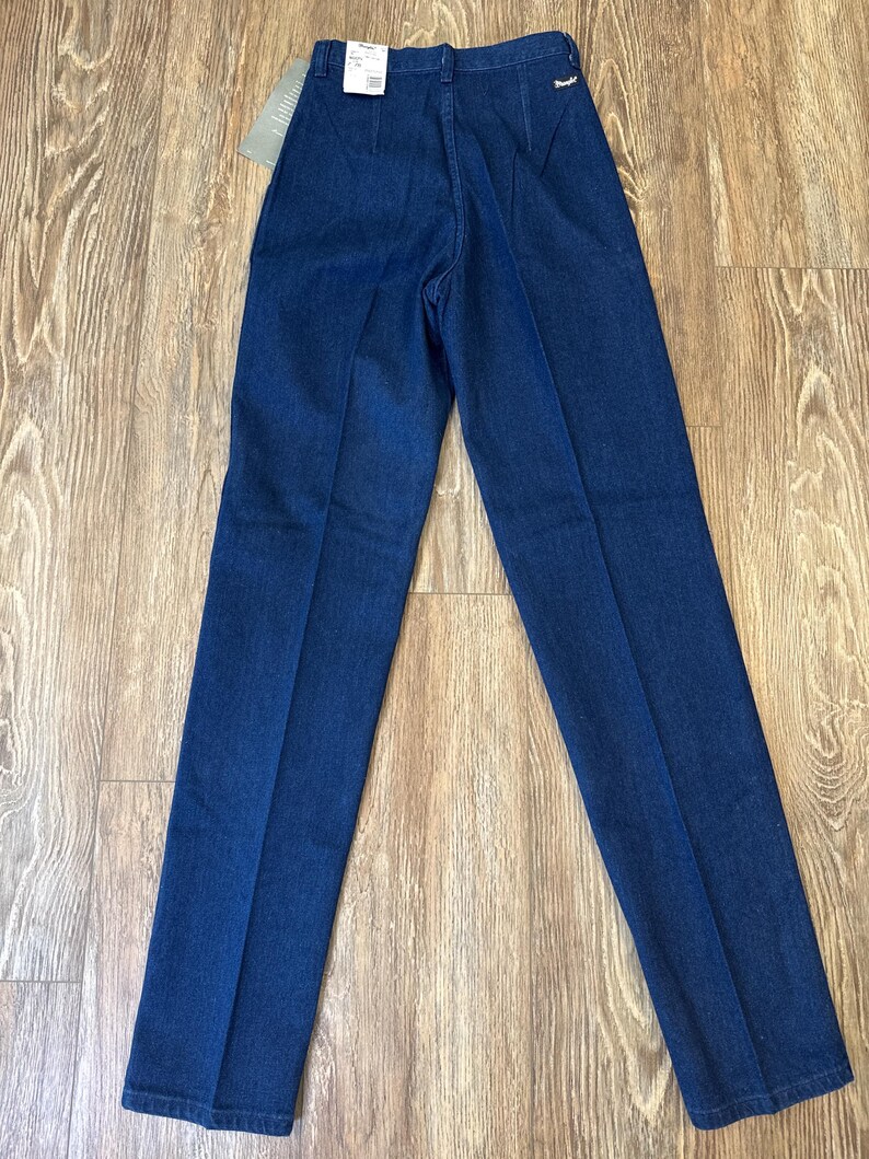 Puede incluir: Pantalones vaqueros azul oscuro con cintura alta y pernera recta. Los pantalones tienen un dise&ntilde;o cl&aacute;sico de cinco bolsillos y una peque&ntilde;a etiqueta de marca en la parte trasera. Una etiqueta de precio est&aacute; adherida a la cintura.