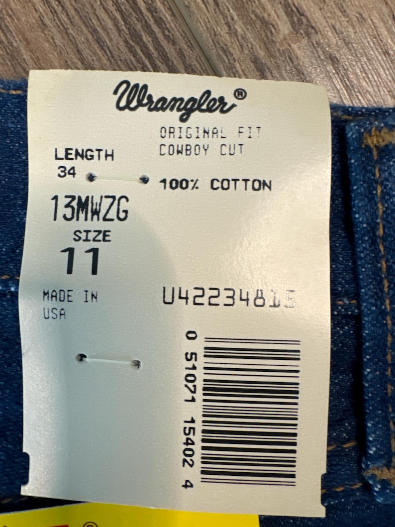 May include: A white tag with black text that reads "Wrangler", "Original Fit Cowboy Cut", "Length 34", "13MWZG Size 11", "100% Cotton", "Made in USA", and "042234815". A barcode is also visible on the tag.