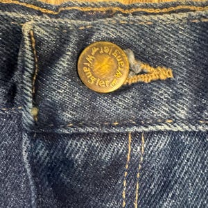 K&ouml;nnte beinhalten: Nahaufnahme eines Abschnitts einer blauen Jeans. Ein Messingknopf mit dem Wort "Wrangler" ist in der Mitte des Bildes zentriert. Der Knopf ist am Bund der Jeans befestigt. Der Denim hat sichtbare N&auml;hte und ein strukturiertes Aussehen.