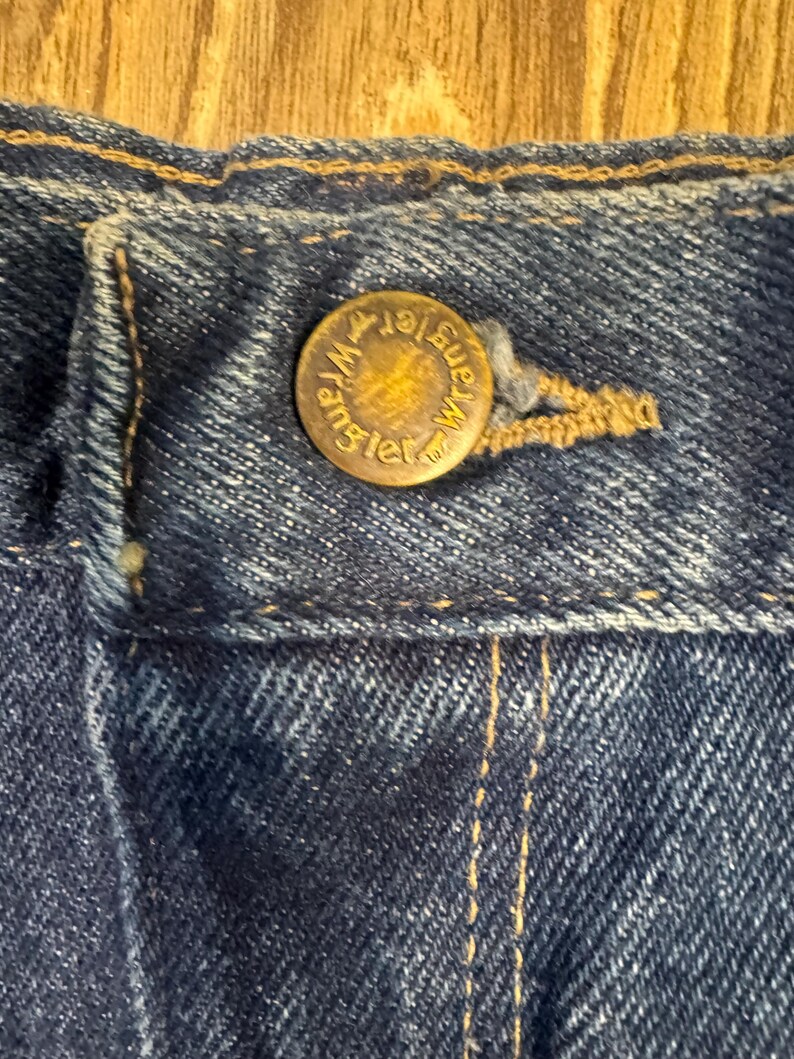 K&ouml;nnte beinhalten: Nahaufnahme eines Abschnitts einer blauen Jeans. Ein Messingknopf mit dem Wort "Wrangler" ist in der Mitte des Bildes zentriert. Der Knopf ist am Bund der Jeans befestigt. Der Denim hat sichtbare N&auml;hte und ein strukturiertes Aussehen.