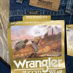 Puede incluir: Primer plano de unos vaqueros Wrangler azules con etiquetas. Las etiquetas dicen "Wrangler Rugged Wear" y "Relaxed Fit". Una etiqueta presenta una ilustraci&oacute;n de personas en una canoa. Los vaqueros son de mezclilla.