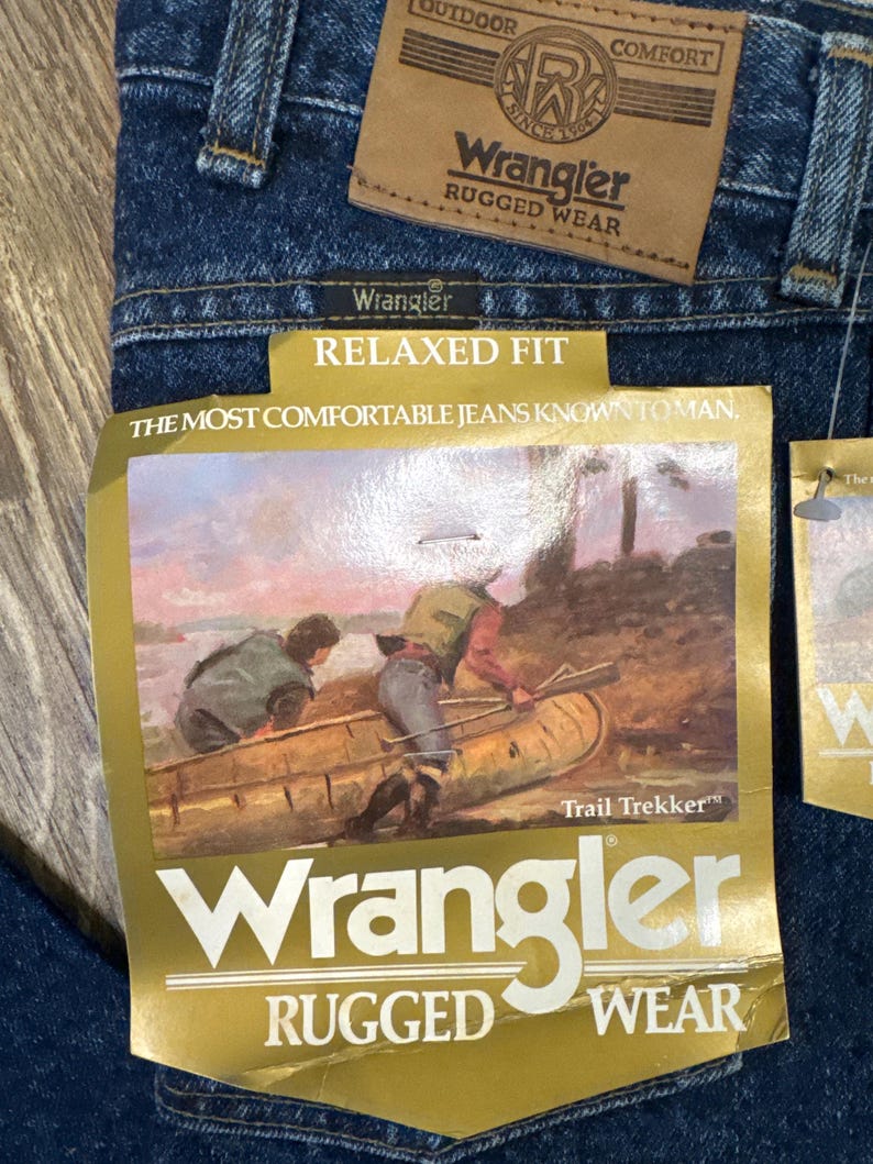 Puede incluir: Primer plano de unos vaqueros Wrangler azules con etiquetas. Las etiquetas dicen "Wrangler Rugged Wear" y "Relaxed Fit". Una etiqueta presenta una ilustraci&oacute;n de personas en una canoa. Los vaqueros son de mezclilla.