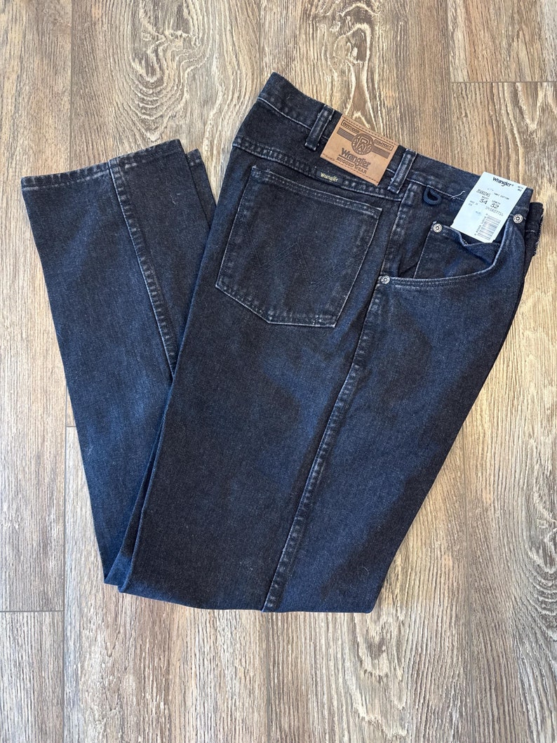 Puede incluir: Un par de pantalones vaqueros negros de denim con una etiqueta Wrangler de cuero marr&oacute;n en el bolsillo trasero. Los vaqueros tienen un corte recto y un dise&ntilde;o cl&aacute;sico de cinco bolsillos.