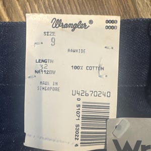 Puede incluir: Una etiqueta de mezclilla Wrangler azul con texto blanco. La etiqueta incluye la talla 9, la longitud 32, y las palabras "Rawhide" y "100% Algod&oacute;n". La etiqueta tambi&eacute;n incluye las palabras "Hecho en Singapur" y un c&oacute;digo de barras con el n&uacute;mero 042670240.
