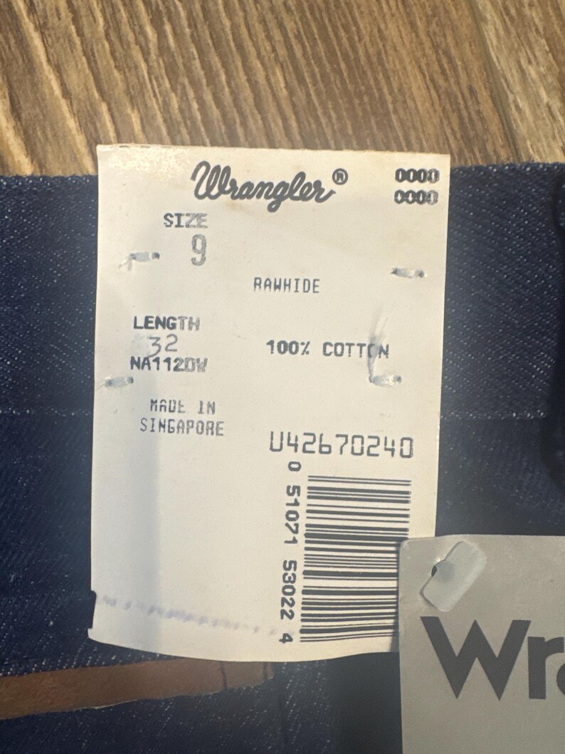 Puede incluir: Una etiqueta de mezclilla Wrangler azul con texto blanco. La etiqueta incluye la talla 9, la longitud 32, y las palabras "Rawhide" y "100% Algod&oacute;n". La etiqueta tambi&eacute;n incluye las palabras "Hecho en Singapur" y un c&oacute;digo de barras con el n&uacute;mero 042670240.
