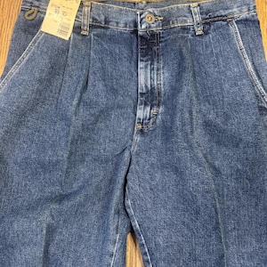 May include: Close-up of blue denim Wrangler jeans with a pleated front. The jeans have a button closure and belt loops. A tag is attached to the jeans with the Wrangler logo and size information.