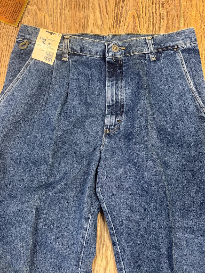 May include: Close-up of blue denim Wrangler jeans with a pleated front. The jeans have a button closure and belt loops. A tag is attached to the jeans with the Wrangler logo and size information.