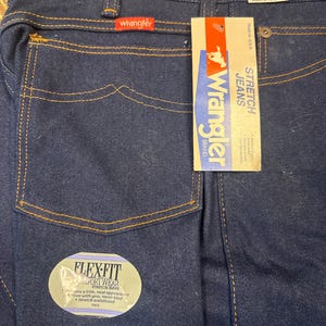 May include: Dark blue Wrangler stretch jeans with a Flex-Fit label. The jeans have a classic five-pocket design with orange stitching. A Wrangler brand tag is attached, and the jeans are size 34 with an 86cm inseam.