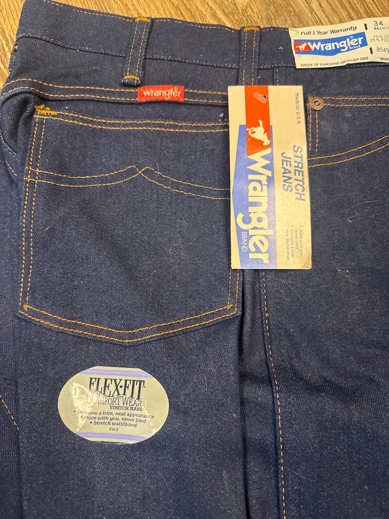 May include: Dark blue Wrangler stretch jeans with a Flex-Fit label. The jeans have a classic five-pocket design with orange stitching. A Wrangler brand tag is attached, and the jeans are size 34 with an 86cm inseam.