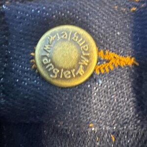 K&ouml;nnte beinhalten: Nahaufnahme eines goldfarbenen Metallknopfes mit dem gepr&auml;gten Text "Wrangler". Der Knopf ist an einem dunkelblauen Jeansstoff befestigt.