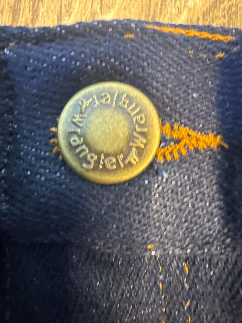 K&ouml;nnte beinhalten: Nahaufnahme eines goldfarbenen Metallknopfes mit dem gepr&auml;gten Text "Wrangler". Der Knopf ist an einem dunkelblauen Jeansstoff befestigt.