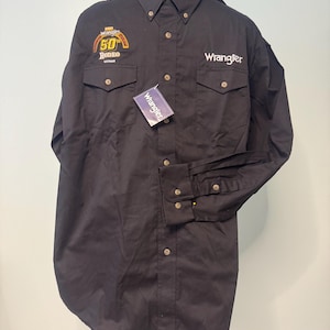 May include: A black long-sleeve button-down shirt featuring a Wrangler logo on the left chest and a 50th Wrangler Rodeo Las Vegas patch on the right chest. The shirt includes two chest pockets with button flaps and a Wrangler brand tag attached.