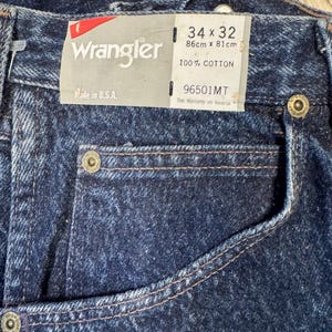 May include: Close-up of dark blue Wrangler jeans with a tag. The tag reads "Wrangler", "Made in U.S.A.", and size 86cm x 81cm. The jeans are 100% cotton.