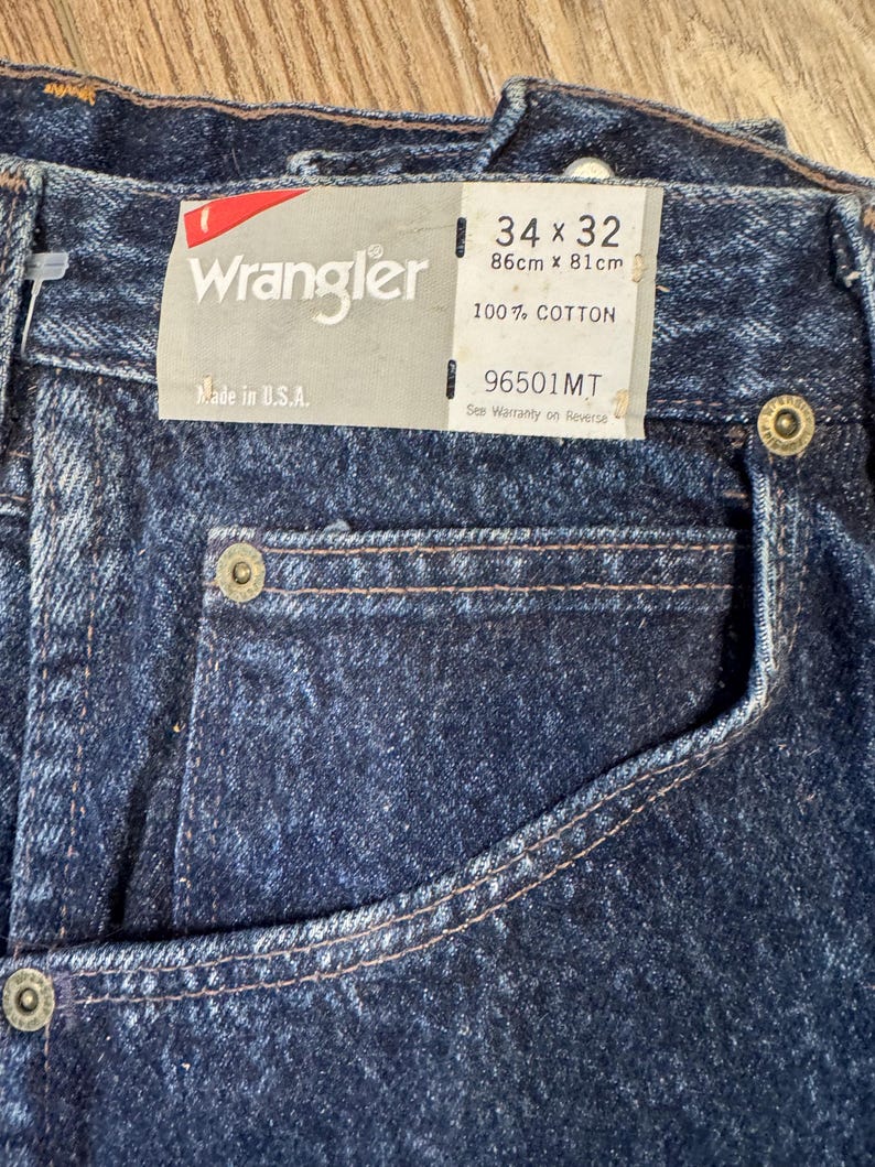 May include: Close-up of dark blue Wrangler jeans with a tag. The tag reads "Wrangler", "Made in U.S.A.", and size 86cm x 81cm. The jeans are 100% cotton.