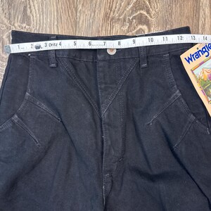 May include: Black denim Wrangler jeans with a tape measure showing a waist measurement of approximately 12 inches. The jeans have a button closure and a classic five-pocket design. A Wrangler tag is visible.