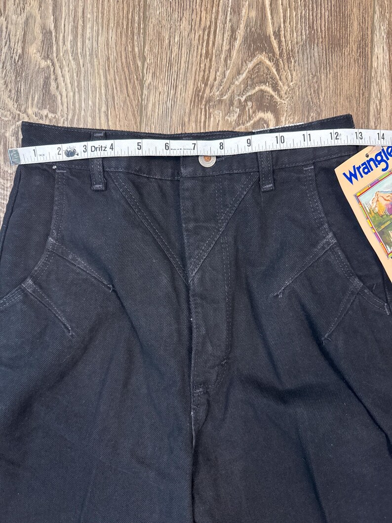 May include: Black denim Wrangler jeans with a tape measure showing a waist measurement of approximately 12 inches. The jeans have a button closure and a classic five-pocket design. A Wrangler tag is visible.